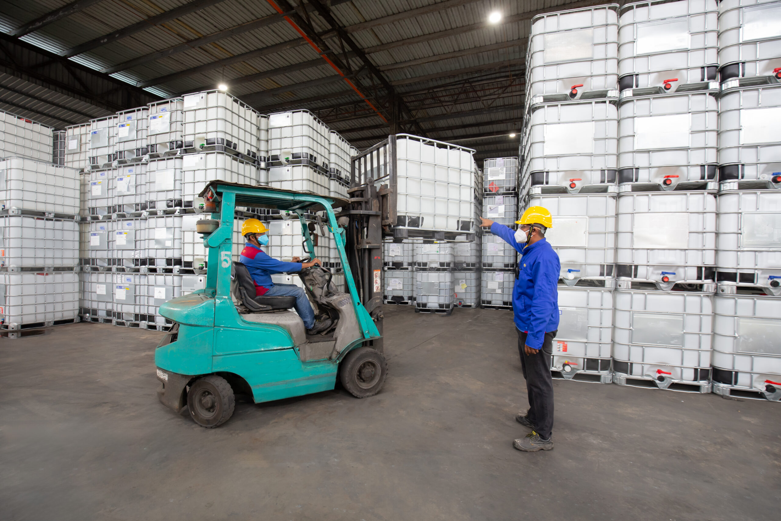 Two workers moving an IBC tote tank with a forklift among stacked containers