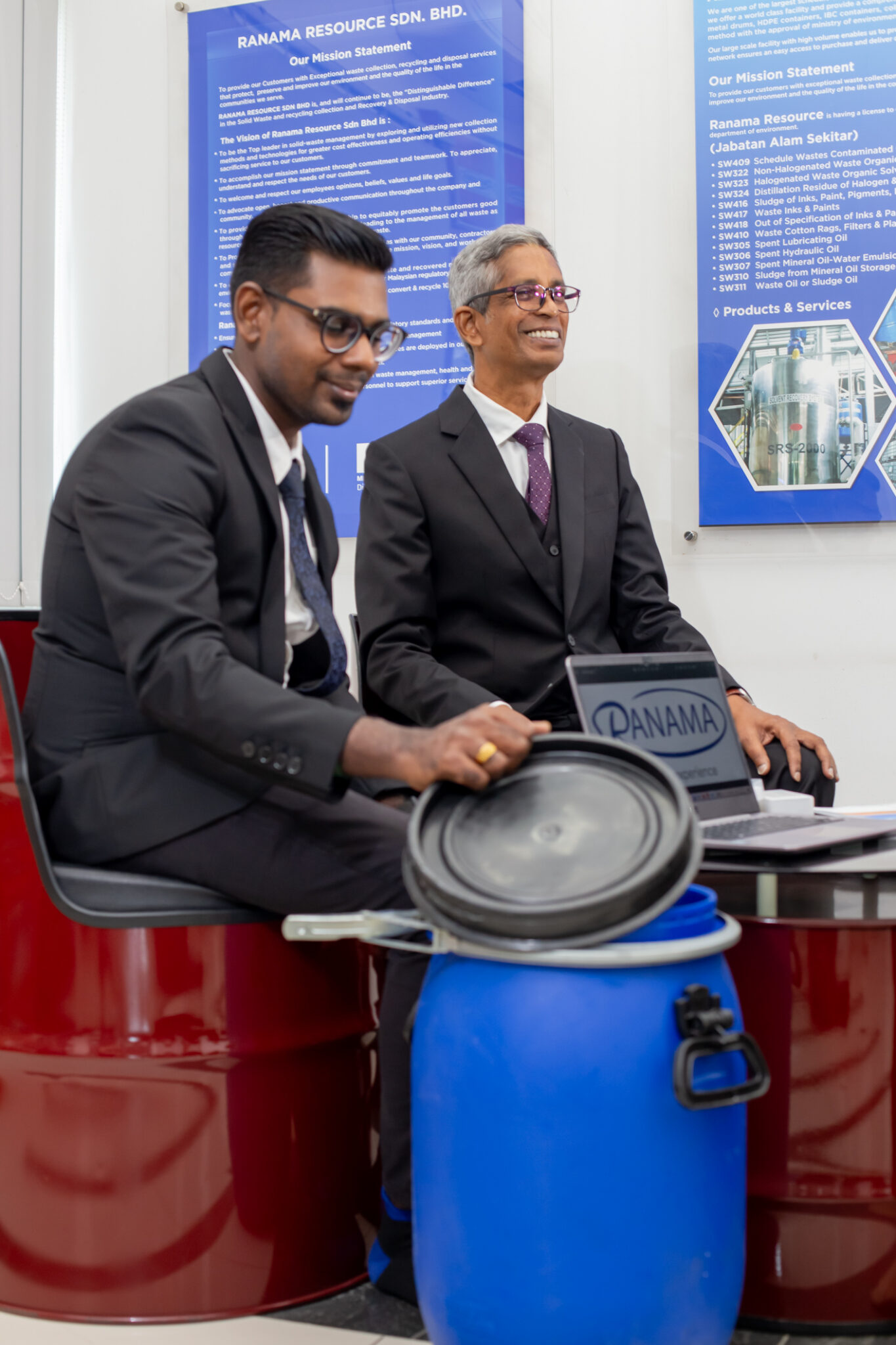 Ranama representatives seated beside industrial drums and a blue container